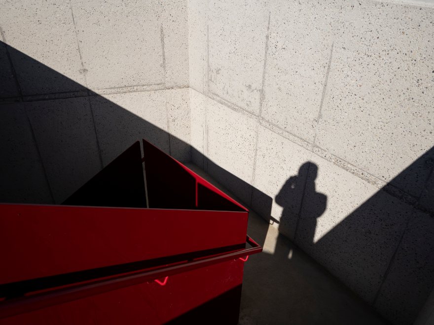 Sunlight and shadow across a concrete stairwell with a red steel handrail, and the shadow of a person on the wall.